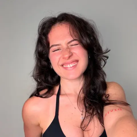 Sarah Mackay Sarah Mackay smiles and squints at the camera while taking a selfie. She has long dark brown hair and is wearing a black sleeveless top; she's standing against a plain white wall.
