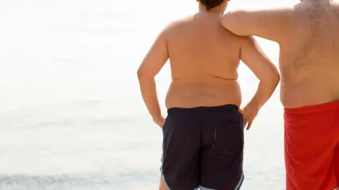 Getty Images A boy and his father stand in their swimming trunks looking at the sea. Both are overweight or obese