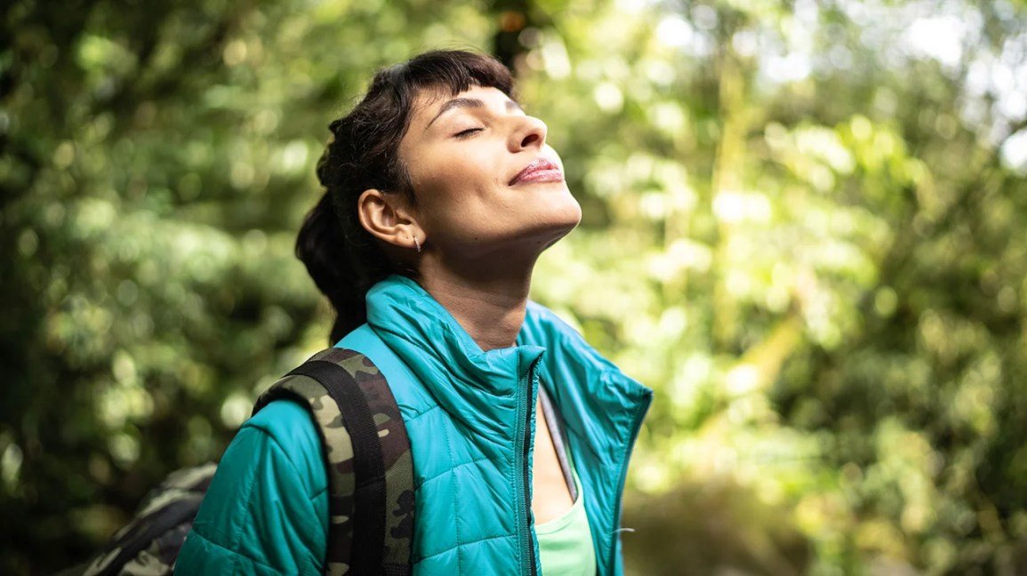 Person wearing hiking clothes and backpack stands in woods smiling with eyes closed
