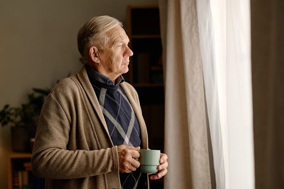 Thoughtful Senior Man Standing by Window with Hot Drink