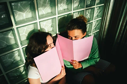 Getty Images Two friends smile at each other from behind pink menus (Credit: Getty Images)