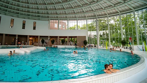 Alamy A large indoor thermal pool under a domed roof with people swimming or resting on the curved edge (Credit: Alamy)